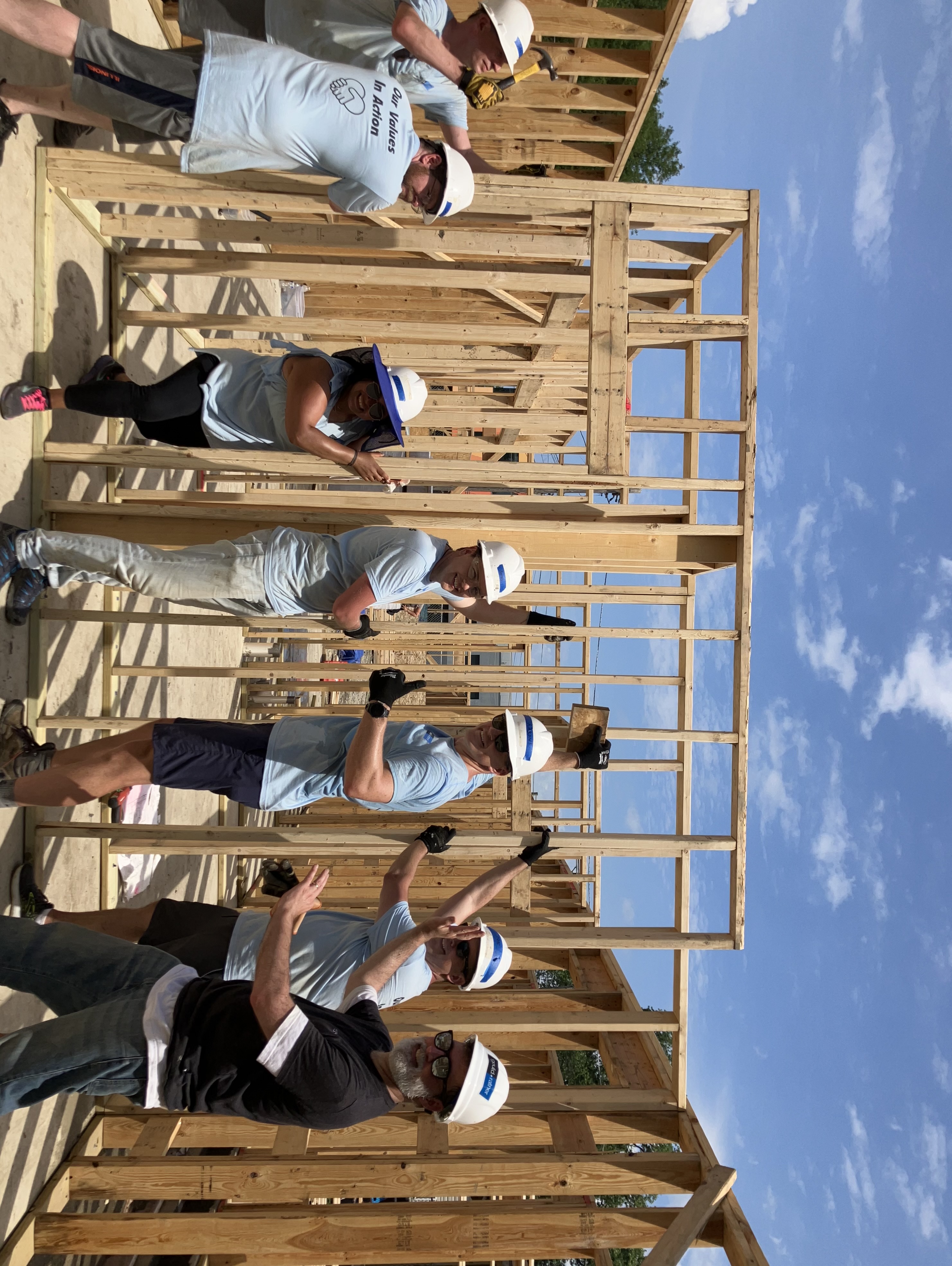 Seven people on a build site posing, holding a framed wall up