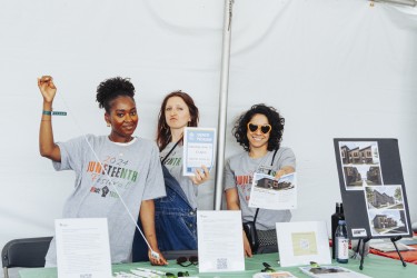 Three women behind a info table holding flyers and a small tape measure