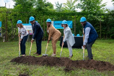 Five people in blue hard hats, each holding a shovel ready to dig into dirt