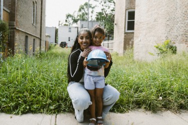 Woman with her daughter, holding a blue hard hat, standing in front of an empty lot